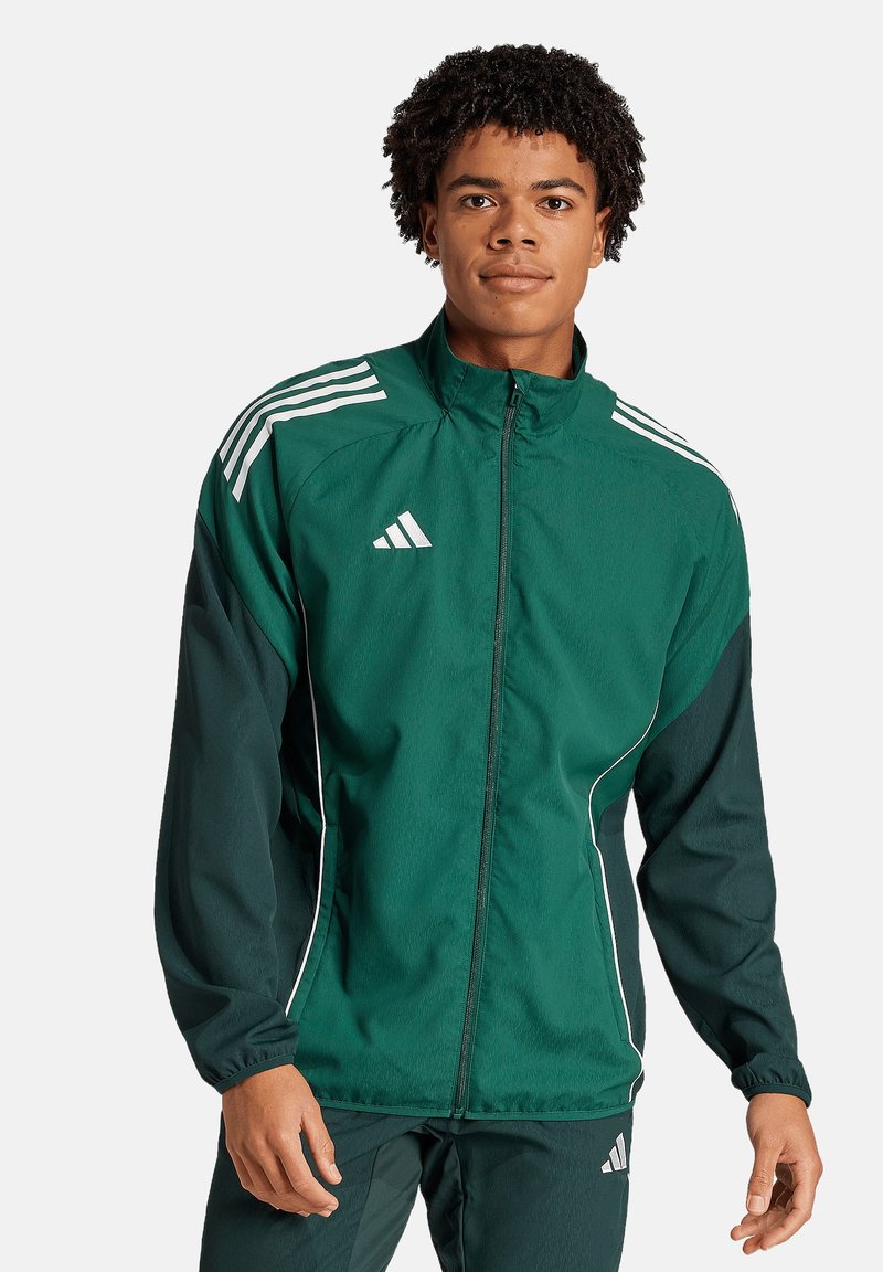 Groene sportjack met contrasterende donkergroene mouwen, witte strepen op de schouders, rits aan de voorkant en elastische manchetten. Bevat het Adidas-logo.