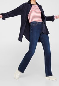 Camicia in velluto a coste blu navy con colletto a punta sopra una maglietta a maniche lunghe rossa e bianca a righe, abbinata a jeans svasati blu scuro e sneakers bianche.