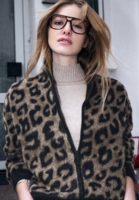 Kardigan w panterkę z miękkiego sztucznego futra, z zamkiem z przodu, czarnym wykończeniem oraz swetrem z wysokim kołnierzem pod spodem, na neutralnym tle.