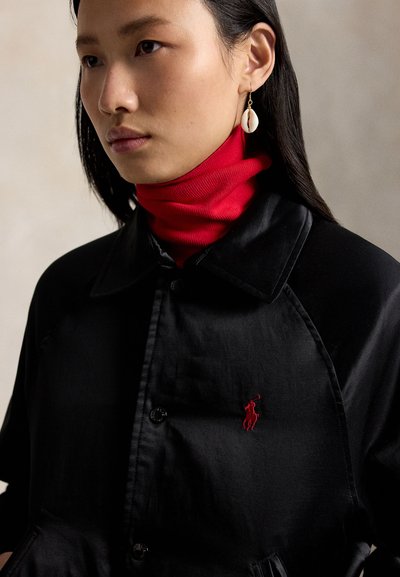 Frau mit langen dunklen Haaren trägt roten Rollkragenpullover unter schwarzer Jacke mit rotem gesticktem Logo und Muschelohring am linken Ohr.