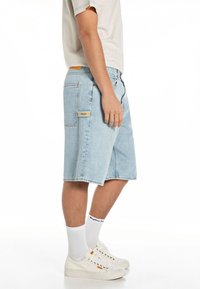 Pantalones cortos de mezclilla azul claro con un ajuste relajado, que cuentan con un bolsillo lateral y un acento amarillo. Combinados con zapatillas blancas y calcetines.