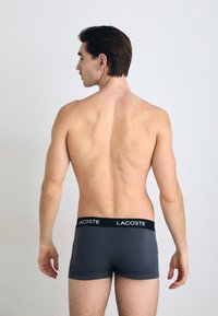 Boxer-briefs gris avec une ceinture noire arborant le logo "LACOSTE", présentés de dos sur un torse nu contre un fond neutre.