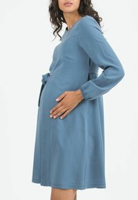 Ein blaues, knielanges, langärmliges Kleid aus weichem Stoff mit rundem Ausschnitt und seitlichen Taschen, entspannter Passform und gerafften Details.