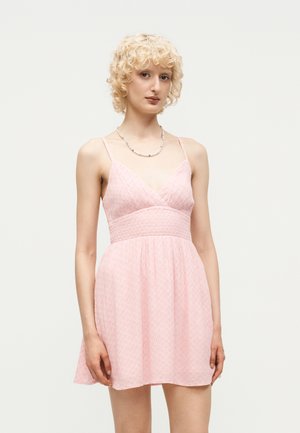 Junge Person mit kurzen lockigen blonden Haaren, trägt ein ärmelloses hellrosa Kleid und eine Perlenkette, steht vor einem einfarbigen Hintergrund.