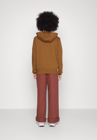 Brun huvtröja med känguruficka och dragsko på huvan, kombinerad med vida, uppvikta byxor i rostfärg och ljusa sneakers.