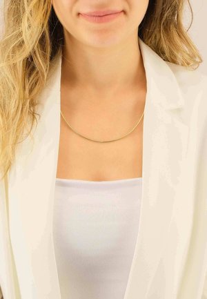 Collana in metallo d'oro con un design liscio, sottile e rotondo. Indossata sopra una maglia bianca, su uno sfondo neutro con distrazioni minime.