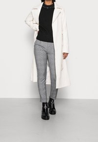 Lång vit trenchcoat, svart ribbad tröja, grå rutig slim-fit byxor, svarta ankelskor med ringdetalj, stående mot en neutral bakgrund.