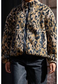 Personne portant une veste zippée en peluche à imprimé léopard avec des poches noires et un jean foncé, se tenant avec les bras détendus le long du corps.