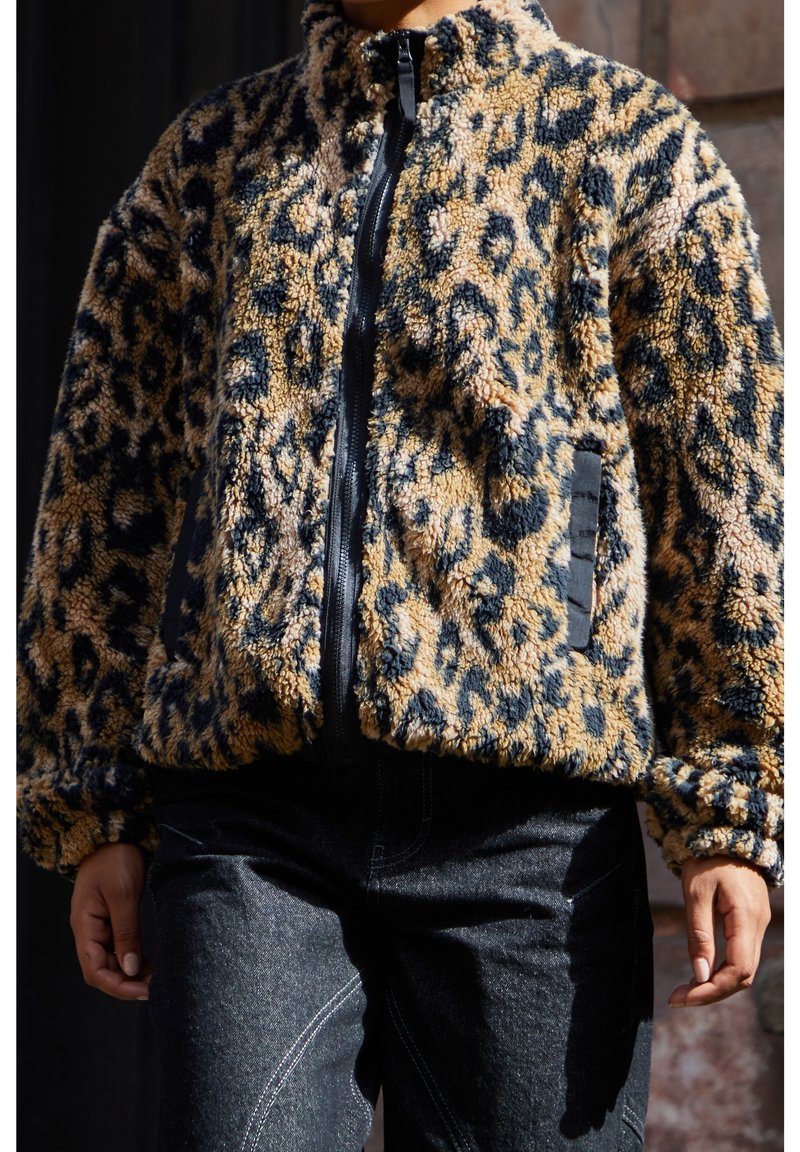 Personne portant une veste zippée en peluche à imprimé léopard avec des poches noires et un jean foncé, se tenant avec les bras détendus le long du corps.