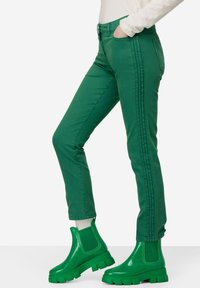 Zielone dżinsowe spodnie z bocznymi paskami, w zestawie z błyszczącymi zielonymi botkami na kostkę. Charakteryzują się dopasowanym krojem i widocznymi detalami ściegów.