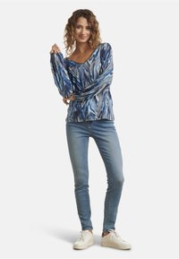 Maglietta a maniche lunghe con motivo astratto blu e grigio, design a V, tessuto leggero, abbinata a jeans in denim aderenti e sneakers bianche.