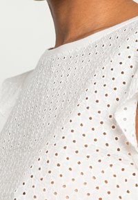 Witte blouse met een getextureerd gaatjesontwerp, voorzien van korte, gerimpelde mouwen en een gladde halslijn. De stof lijkt lichtgewicht en ademend te zijn.