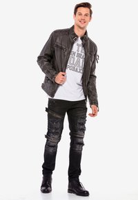 Schwarze Lederjacke mit gestepptenDetails, weißes Grafik-T-Shirt, ausgebeulte schwarze Jeans und schwarze Schnürstiefel. Mann steht lächelnd.