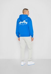 Modra kapuca s belim grafičnim napisom "YOUTH", sproščen kroj. Skombinirana z svetlo sivo cargo hlačami in belimi športnimi copati, kar prikazuje sproščen stil.