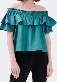 Blouse teal à encolure dégagée avec des détails de volants superposés, fabriquée dans un tissu lisse, assortie à un pantalon noir. Accent collier en chaîne dorée.