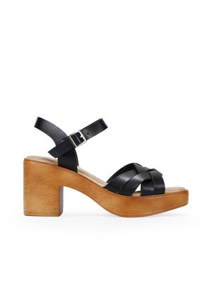 Sandal de cuero negro con diseño de tiras cruzadas, tacón ancho de madera y correa en el tobillo con hebilla. Textura suave y acabado natural de madera.