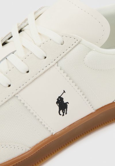 Λευκό αθλητικό παπούτσι με λευκά κορδόνια, μπεζ σουέντ λεπτομέρειες, καουτσούκ σόλα και μαύρο λογότυπο παίκτη πόλο στην πλαϊνή επιφάνεια.
