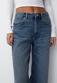 Kvinna som bär högmidjade blå denimjeans och en vit långärmad kort topp, står med händerna avslappnat längs sidorna.