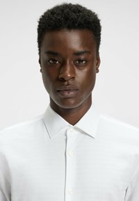 Chemise blanche à boutons, en tissu texturé et à col pointu, affichant une coupe ajustée et des boutons contrastés, portée par un mannequin.