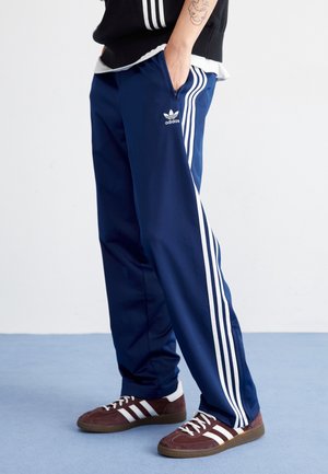 Personne portant un pantalon de jogging Adidas bleu avec des bandes blanches et des baskets Adidas marron, debout les mains dans les poches.