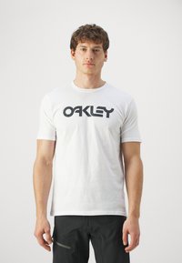 Biała bawełniana koszulka z krótkim rękawem, na której znajduje się duże czarne logo "OAKLEY" z przodu. Posiada okrągły dekolt.