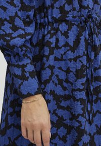 Blaues und schwarzes gemustertes Kleid aus strukturierter Stoff. Es hat einen Kordelzug in der Taille und einen gekrempelten Ärmel. Am Handgelenk wird ein goldener Armreif getragen.