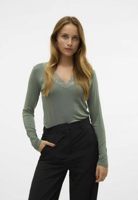 Top de manga larga y escote en V en un suave tejido verde con detalles de encaje; combinado con pantalones negros de cintura alta con bolsillos laterales.