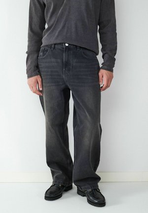 Schwarze Denim-Jeans mit weiter Passform, ausgestattet mit Fronttaschen und dezentem Verblassen. Kombiniert mit einem dunkelgrauen Langarmshirt und schwarzen Schuhen.