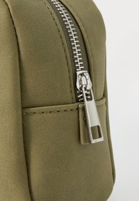 Pochette vert olive fabriquée en matériau lisse, dotée d'une fermeture éclair argentée avec un tirette rectangulaire et des détails de couture subtils.