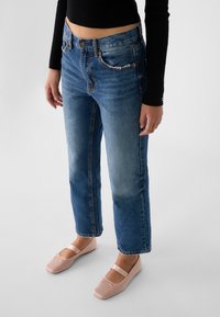 Vaqueros de mezclilla en un tono azul claro, de cintura alta, corte recto y ligeros desgastes. Combinados con bailarinas de tiras en tono rosa pálido.