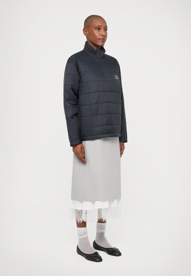 Person im schwarzen gesteppten Pullover, hellgrauem Midikleid mit weißem Spitzenrand, transparenten grauen Socken und schwarzen Ballerinas vor einem weißen Hintergrund.