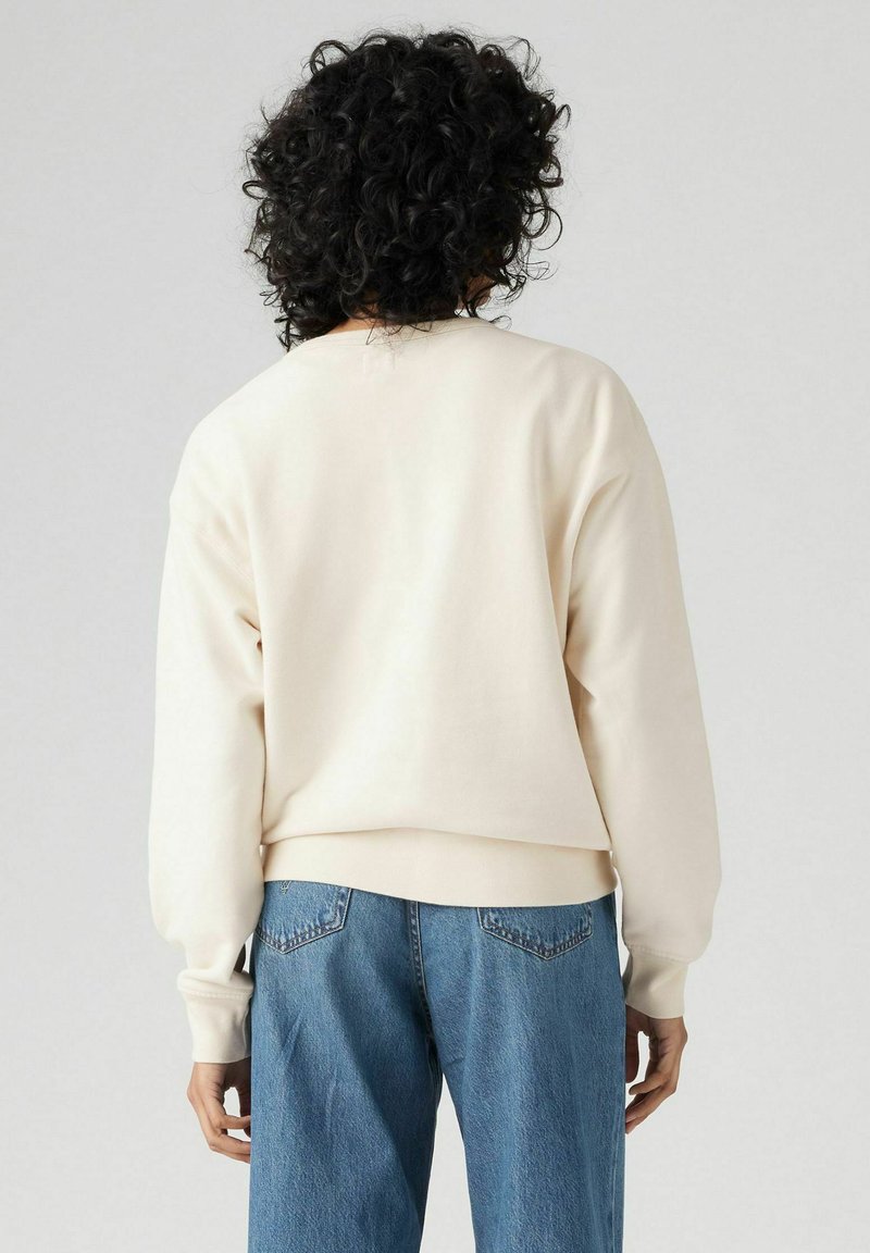 Cremefarbenes Sweatshirt mit überschnittenen Schultern und gerippten Bündchen, kombiniert mit hellblauen Jeans. Weiche Textur und entspannter Schnitt.