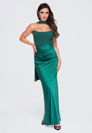 Femme aux longs cheveux foncés portant une robe en satin vert émeraude sans manches avec un corsage ajusté et un col en foulard, posant avec une main sur la hanche.