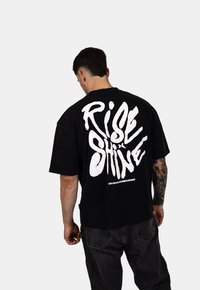 Czarny bawełniany t-shirt z dużym białym nadrukiem tekstu "RISE n SHINE" na plecach. Krótkie rękawy i luźny krój. Zawiera branding współpracy.