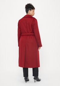 Casaco vermelho longo, feito de tecido macio, com uma cintura cingida por um cinto, colarinho notched e corte reto, combinado com calças pretas e saltos altos.