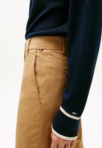 Chinos beiges avec une texture lisse, arborant un petit patch logo. Pull bleu foncé avec une texture côtelée et des poignets blancs contrastants.