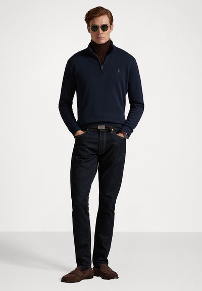 Mørk marineblå half-zip genser med en jevn tekstur; kombinert med mørke jeans og brune loafere. Tilbehøret inkluderer runde solbriller.