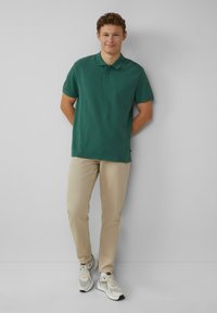 Zielona koszulka polo z trzema guzikami, połączona z beżowymi spodniami i szarymi sneakersami. Casualowy fason i gładka faktura materiału.