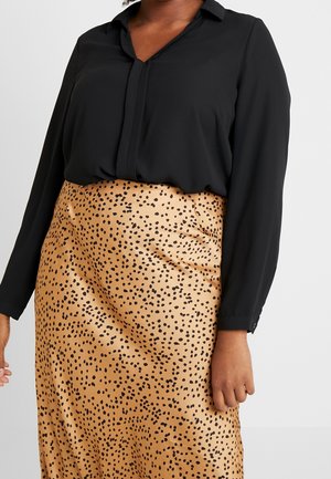 Nainen pukeutuneena pitkähihainen musta paita, joka on tummanruskean hameen sisään sujautettu ja jossa on epäsäännöllisiä mustia pilkkuja, kuvattu olkapäistä polviin.