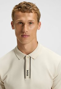 Cremfarbendes Poloshirt mit weicher Textur, ausgestattet mit einer Knopfleiste mit zwei Knöpfen und einem schwarz-weiß gestreiften Kragen mit "BOSS"-Logo.