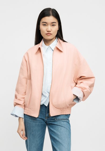 Γυναίκα που φοράει ένα ροζ ανοιχτό μπουφάν με φερμουάρ πάνω από μια μπλε και λευκή ριγέ μπλούζα και μπλε τζιν, στέκεται με το ένα χέρι στην τσέπη.