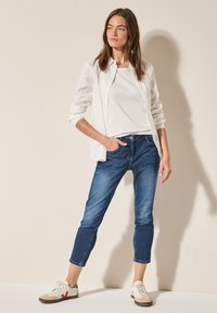 Camisa blanca de botones sobre una blusa blanca, combinada con jeans ajustados en azul oscuro y zapatillas beige con acentos rojos. Diseño sencillo y casual.