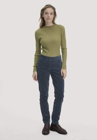 Vrouw die staat in een olijfgroen ribbeltrui, marineblauwe corduroy broek en donkerbruine instappers tegen een effen lichte achtergrond.