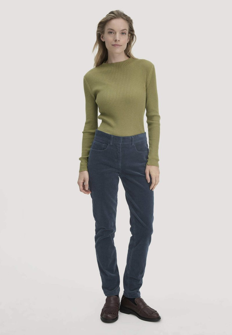 Vrouw die staat in een olijfgroen ribbeltrui, marineblauwe corduroy broek en donkerbruine instappers tegen een effen lichte achtergrond.