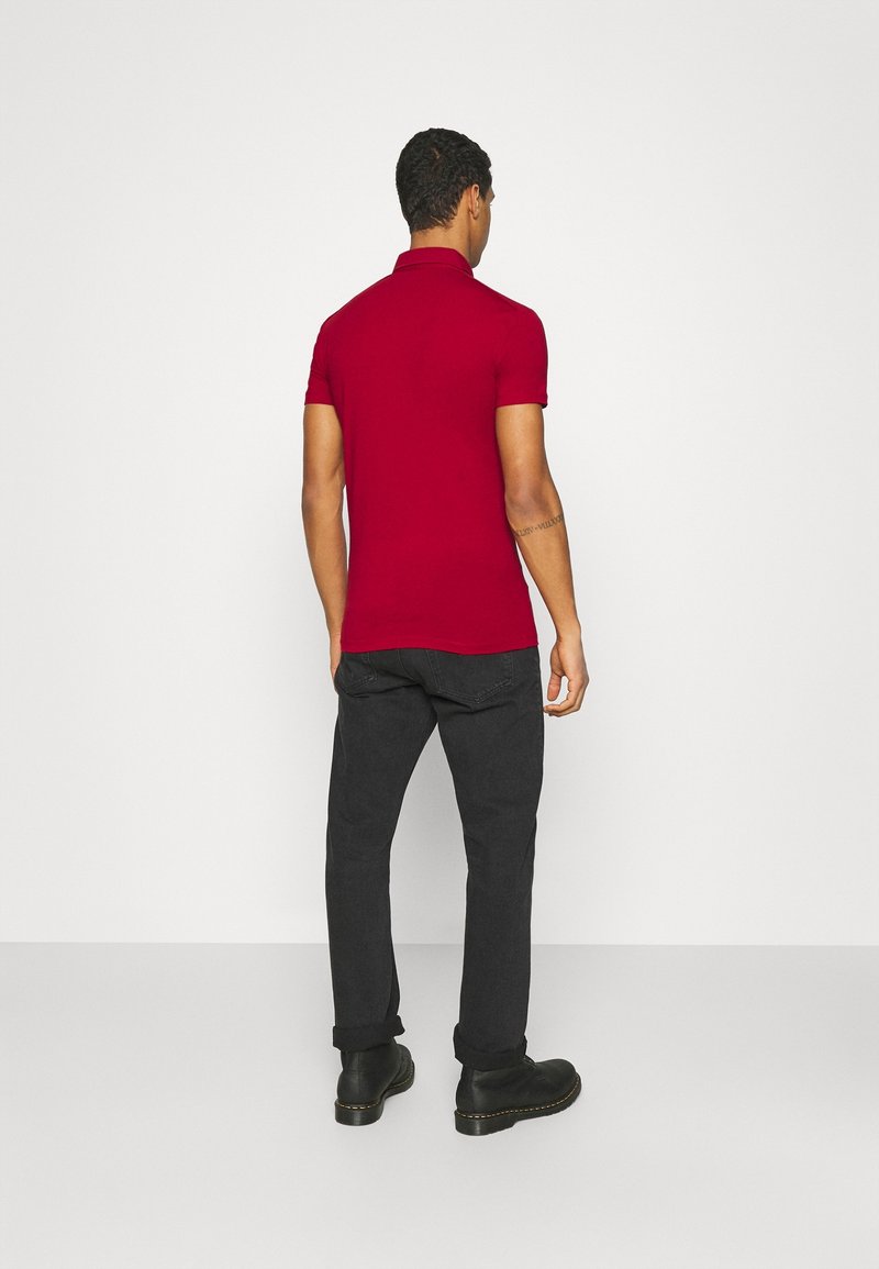 Polo rouge à col court, associé à un pantalon gris foncé et des bottes noires. Design simple, coupe ajustée, sans motifs visibles.