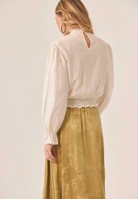 Blusa blanca texturizada con mangas largas, un escote en forma de lágrima en la parte posterior, y cintura fruncida, combinada con una falda larga con un patrón en tonos dorados y verdes.