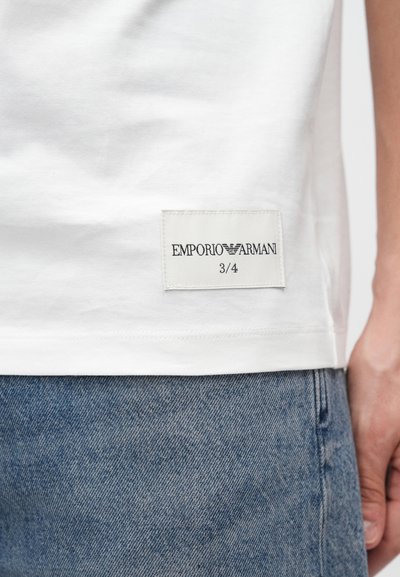 Λευκό βαμβακερό T-shirt με ραμμένη ετικέτα που γράφει "EMPORIO ARMANI 3/4" στη ραφή του πλάγιου μέρους, σε συνδυασμό με μπλε τζιν παντελόνι.