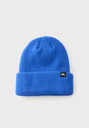 Bonnet en laine bleu tricoté avec un revers plié et une petite étiquette noire avec un logo sur le côté, présenté sur un fond gris clair.