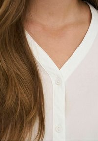 Chemisier blanc avec un décolleté en V, comportant deux boutons, un tissu texturé et des cheveux visibles drapés sur l'épaule.