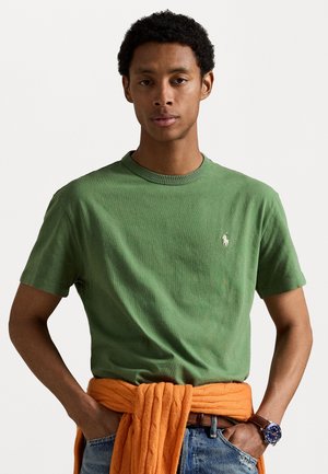 Camiseta de algodón verde de cuello redondo con mangas cortas y un pequeño logotipo bordado, combinada con un suéter de punto trenzado naranja atado en la cintura.